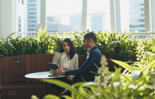 A man and woman are seated at a table, engaged with a tablet in front of them, sharing ideas and collaborating.