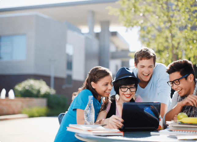 A group of four young people sitting outside, focused on a laptop, collaborating and enjoying the fresh air and sunshine.