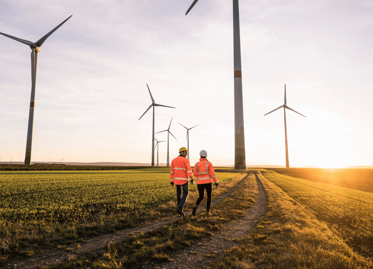 Two people wearing safety vests stroll past towering wind turbines, highlighting renewable energy efforts.