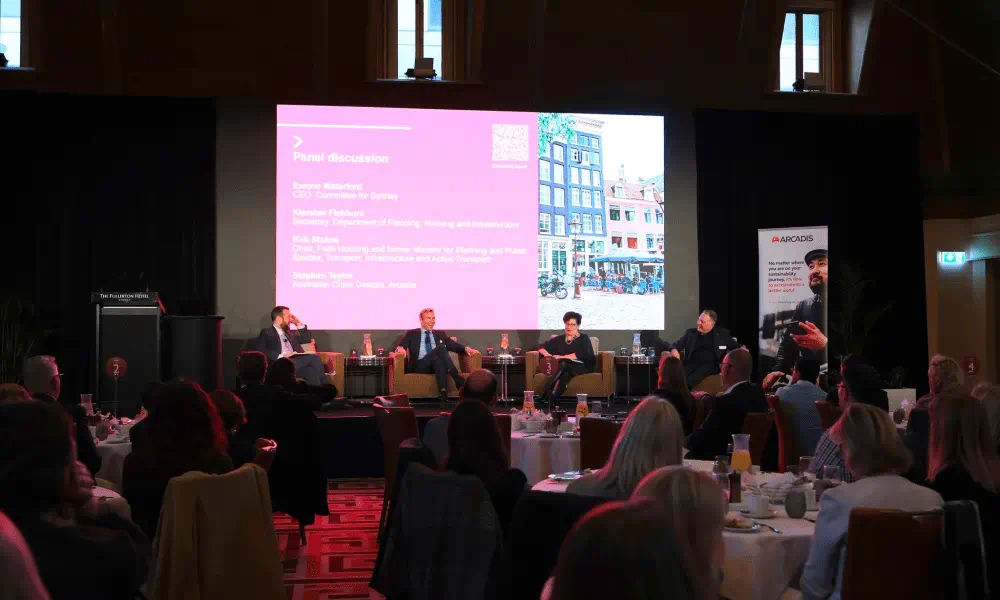 A diverse group of individuals seated at tables, engaged in discussion, with a large screen displaying information in the background.