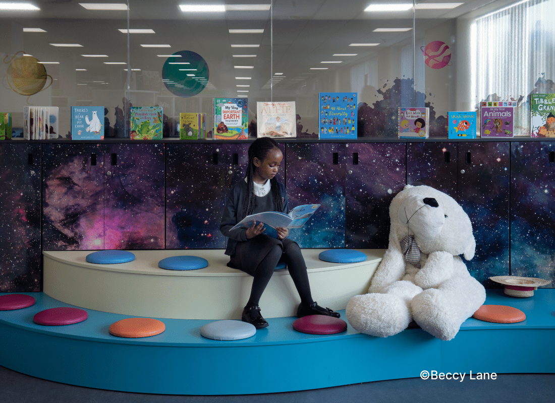 A girl sits at a blue and white round table, smiling beside a plush stuffed bear.