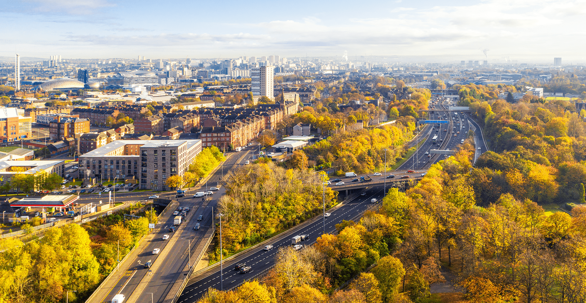 A city skyline featuring tall buildings and lush trees in the foreground against a clear blue sky.