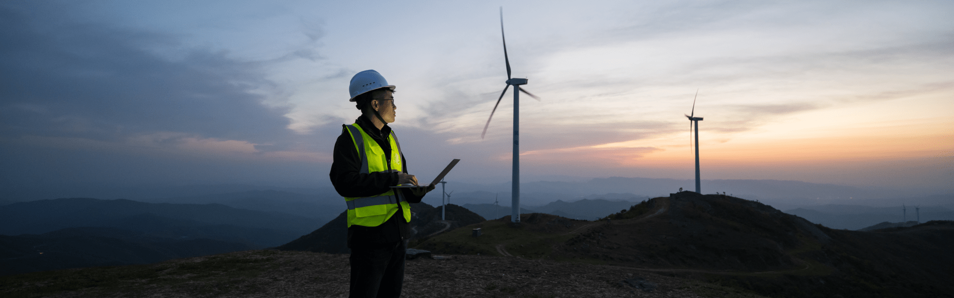 An man in a hard hat overlooking wind turbines atop a mountain.