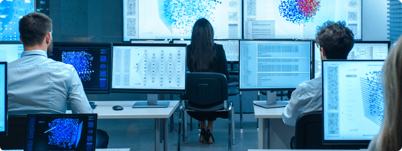 People collaborating at computers in a workspace featuring an array of screens, emphasizing a tech-driven environment.
