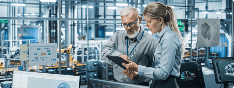 Two factory workers reviewing information on a tablet, collaborating on tasks in a manufacturing environment.