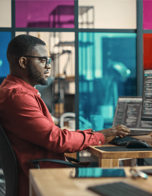 A man seated at a desk, working on two computers, focused on his tasks in a modern office environment.