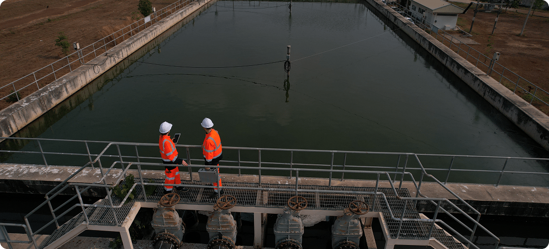 Two men in orange vests stand on a bridge, observing a waterway below them.