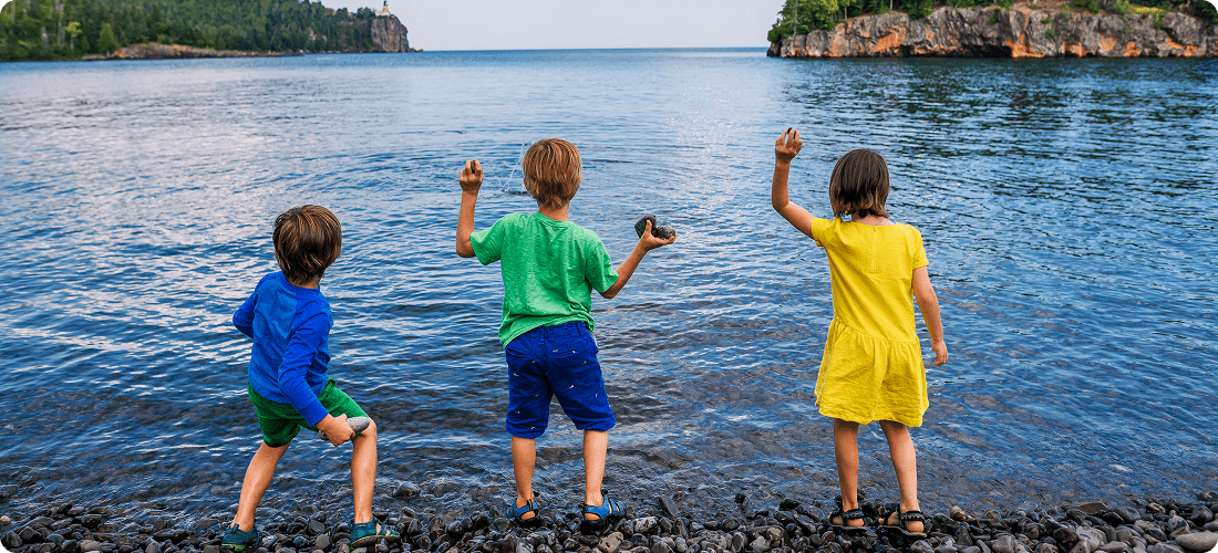 Three children stand on the shore of a lake, gazing at the water with trees and mountains in the background.