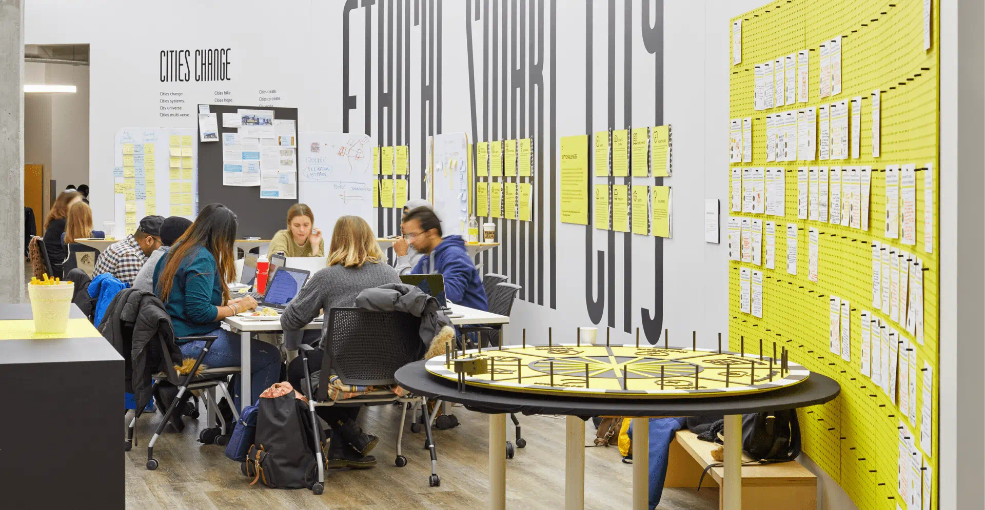  A group of individuals seated at tables, surrounded by yellow sticky notes, engaged in discussion within a bright room.