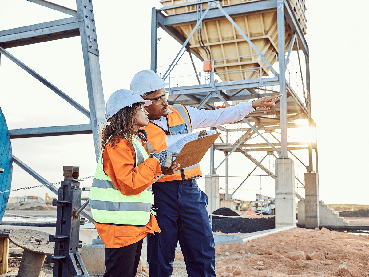Two individuals in hard hats and safety vests stand before a construction site, discussing the ongoing project.