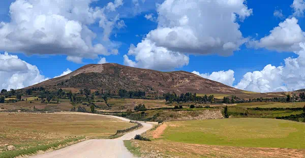 A dirt road winds through a grassy field, leading to a mountain range in the background under a clear blue sky.
