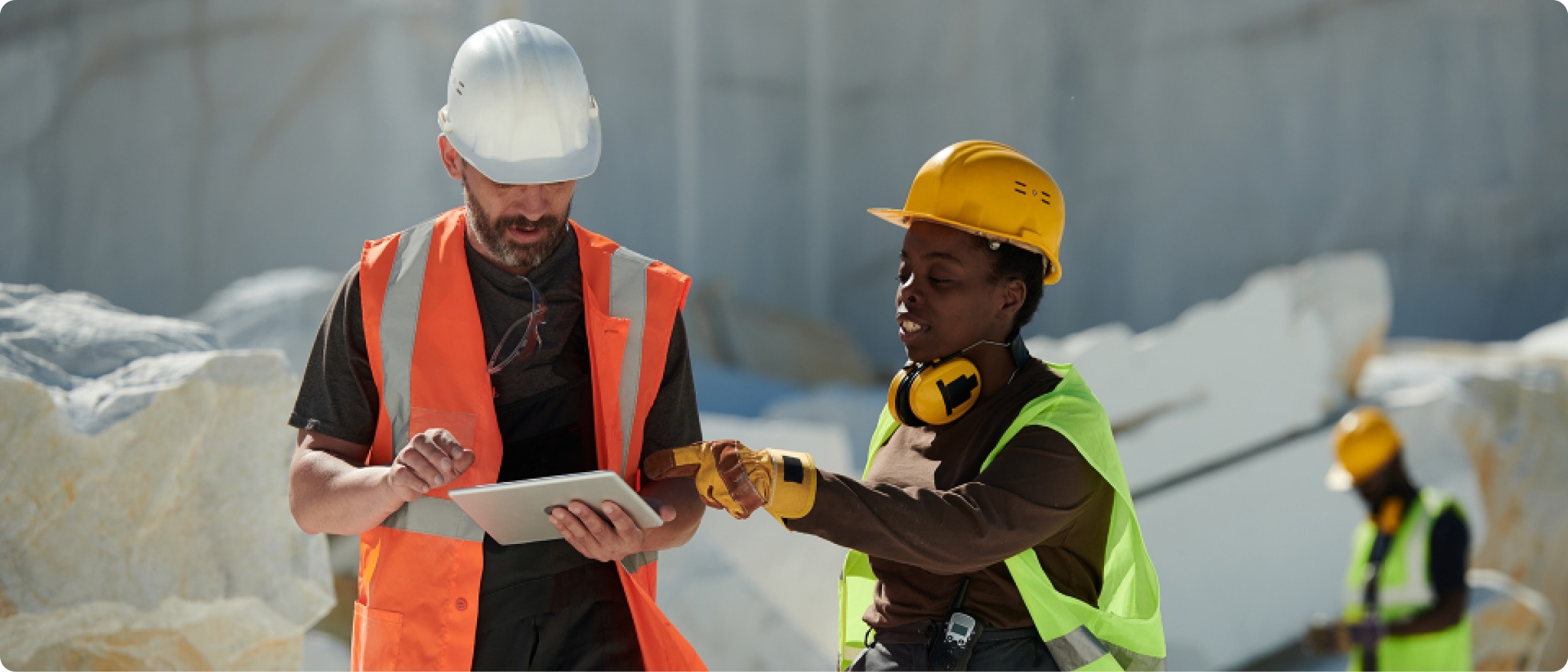 Two construction workers wearing safety vests and hard hats review information on a tablet at an outdoor work site, with large stone blocks and another worker visible in the background.