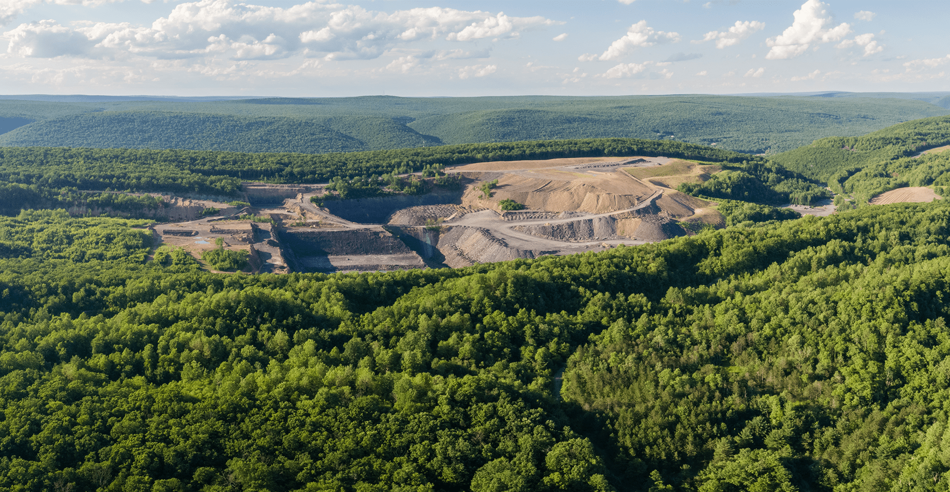 A wide aerial view of a large open-pit mining site surrounded by expansive green forest and rolling hills under a partly cloudy sky.