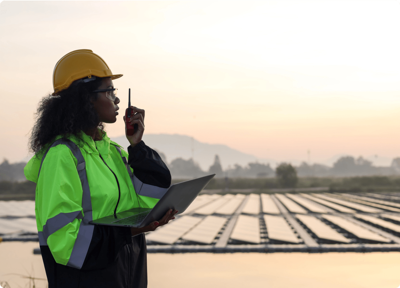 A woman wearing a hard hat and safety vest holds a laptop and radio, standing in front of a vast field.