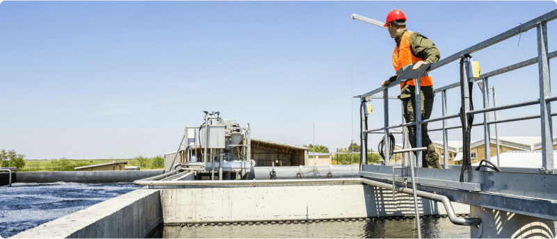 A man wearing an orange vest gazes at a water treatment plant from a platform, highlighting his role in environmental management.