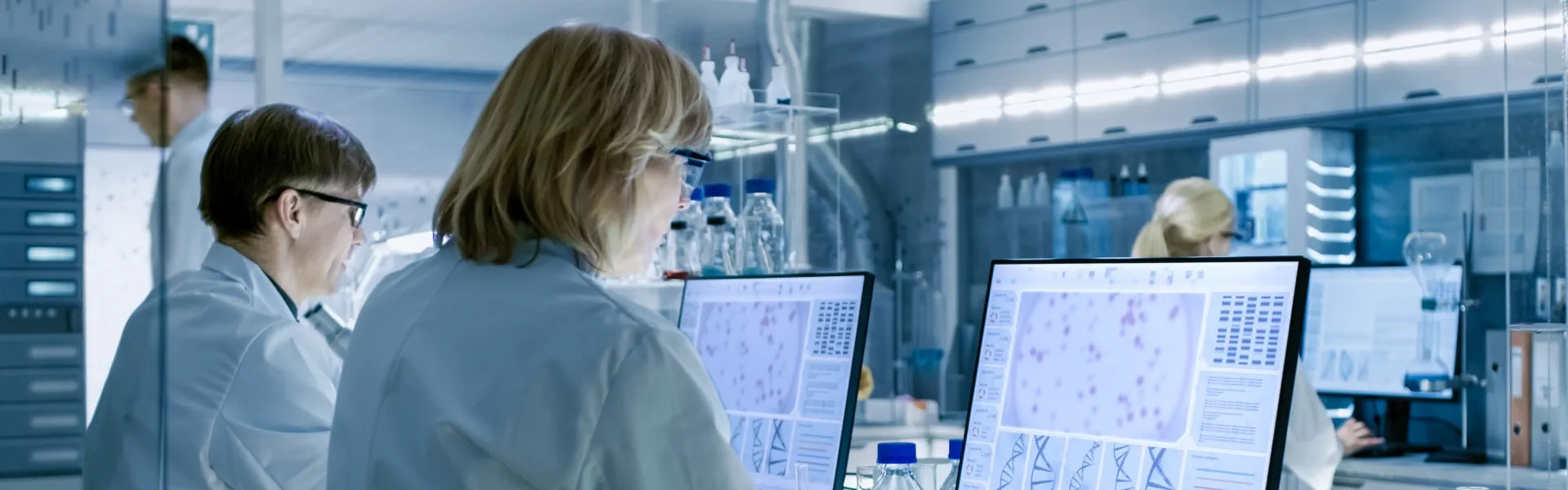 Two individuals in lab coats collaborating on computers in a laboratory setting, focused on their tasks.