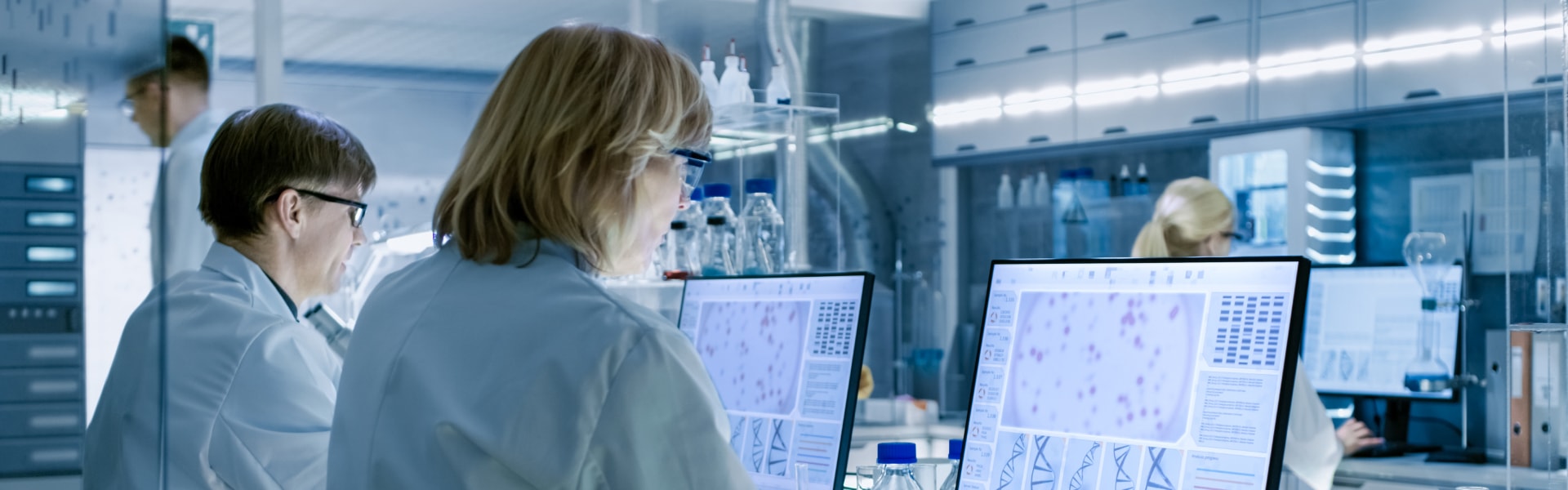 Two individuals in lab coats collaborating on computers in a laboratory setting, focused on their tasks.