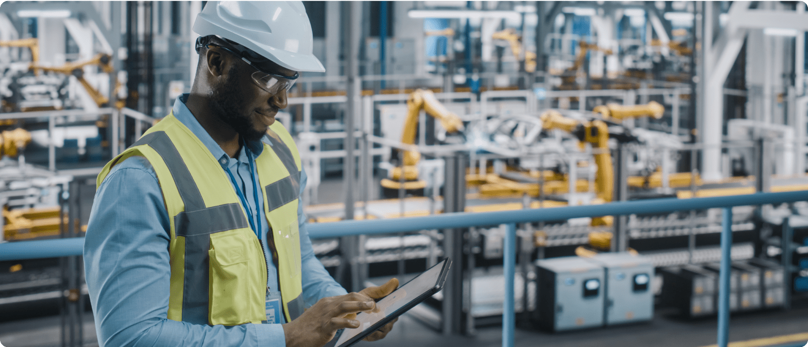 A man wearing a hard hat and safety vest stands confidently in front of a factory building.