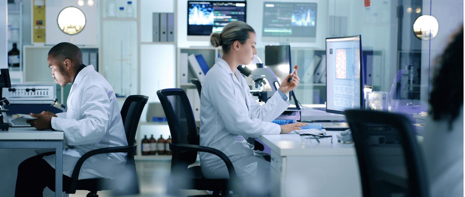A diverse group of individuals in lab coats collaborating on computers in a modern laboratory setting.