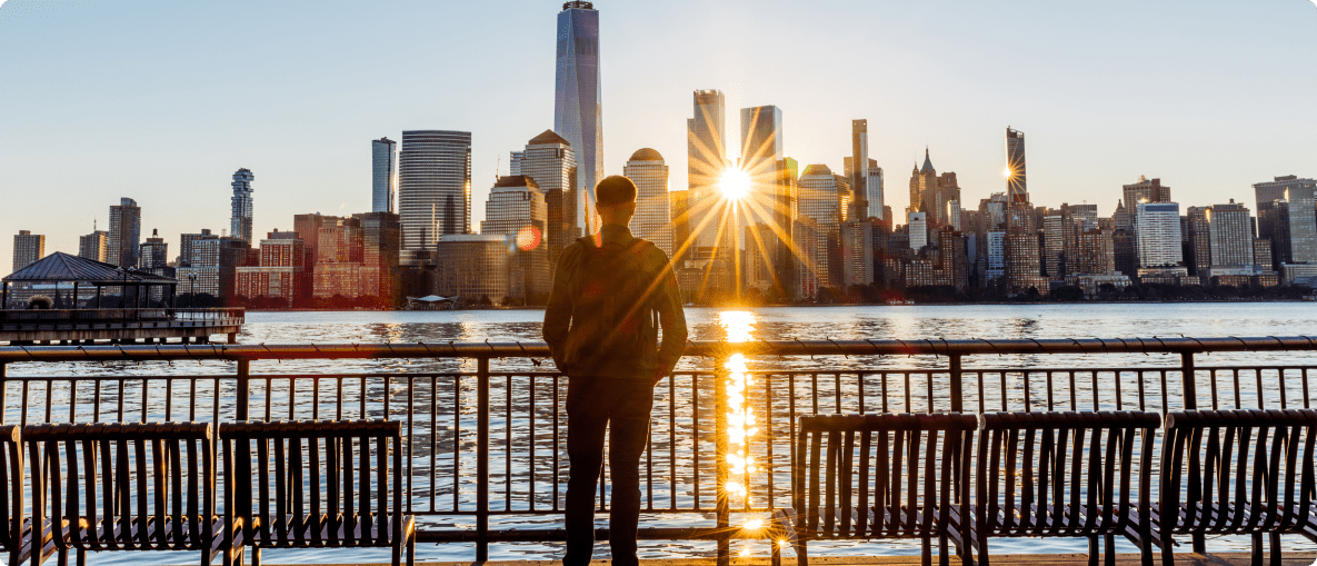 A man stands on a bench, gazing at the city skyline with tall buildings and a clear blue sky in the background.