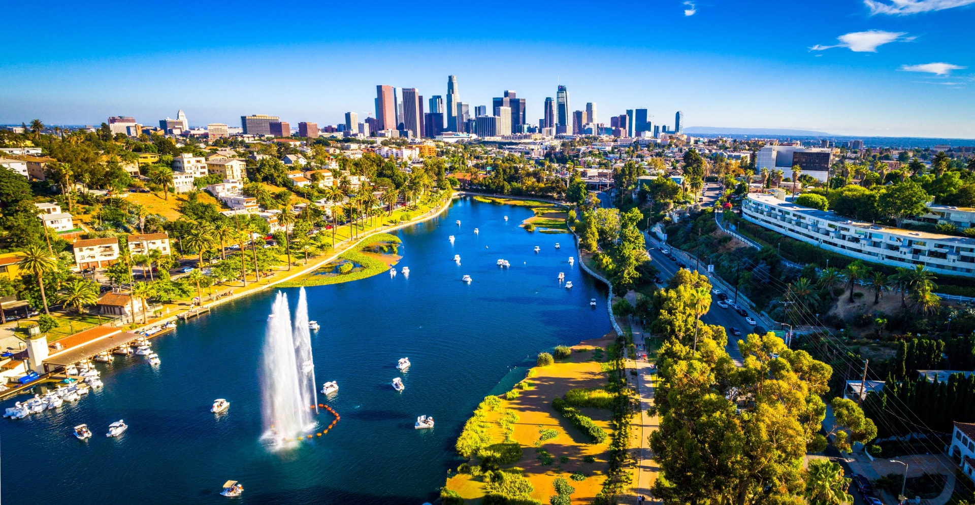 An aerial view of Los Angeles, California’s Echo Park with a water fountain spraying in a lake and the city skyline in the background.