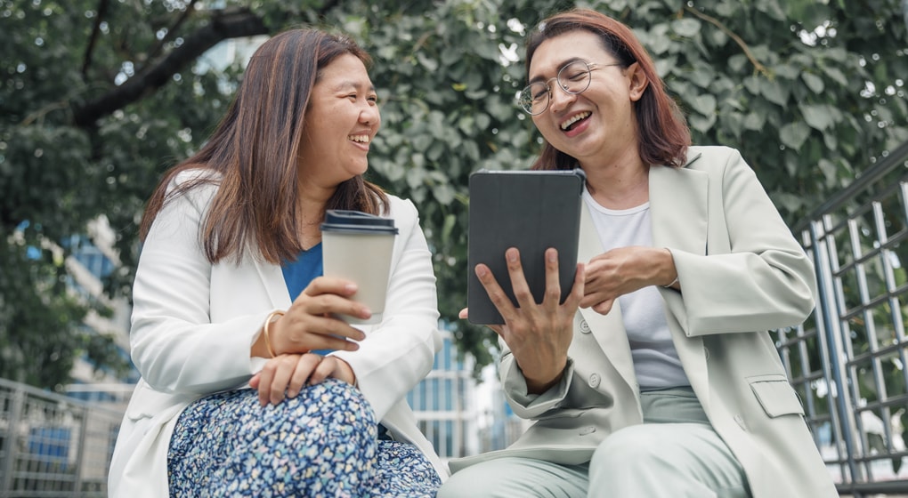 Two women sitting on a bench, looking at a tablet computer together.