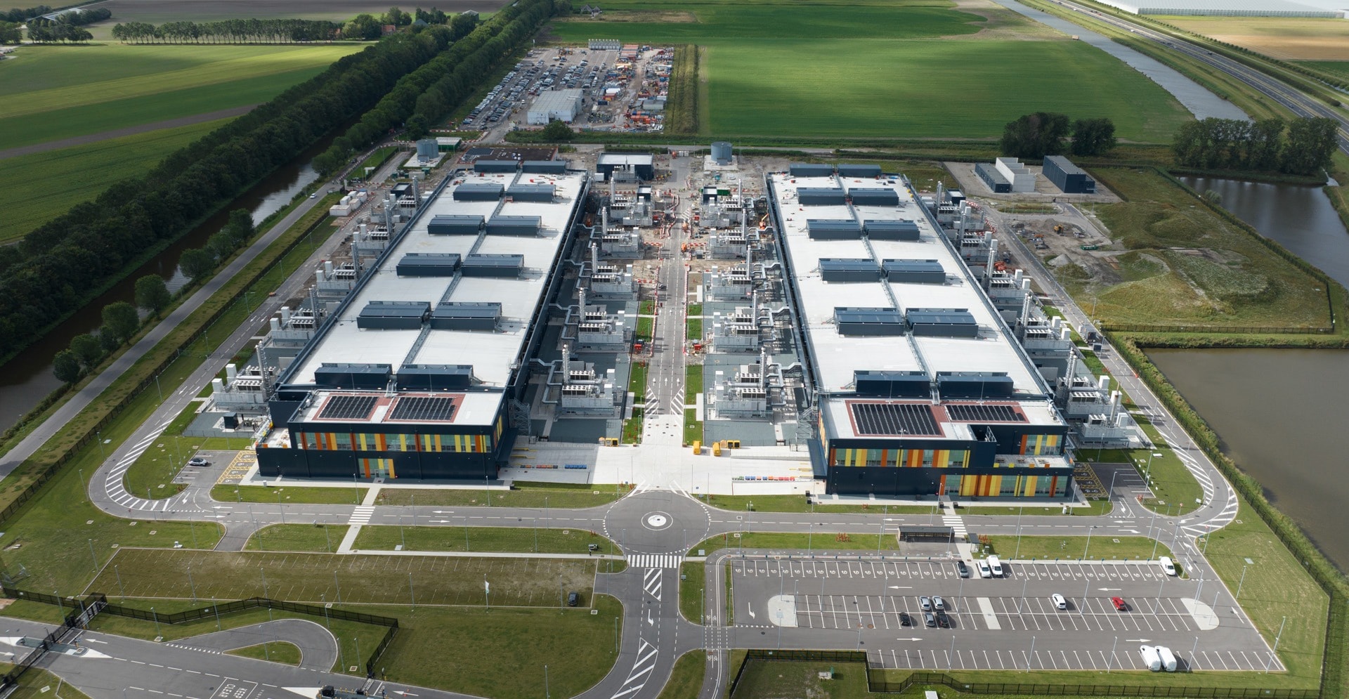 Aerial view of a large, modern industrial complex with two long, rectangular buildings surrounded by parking areas, service roads, and green fields.