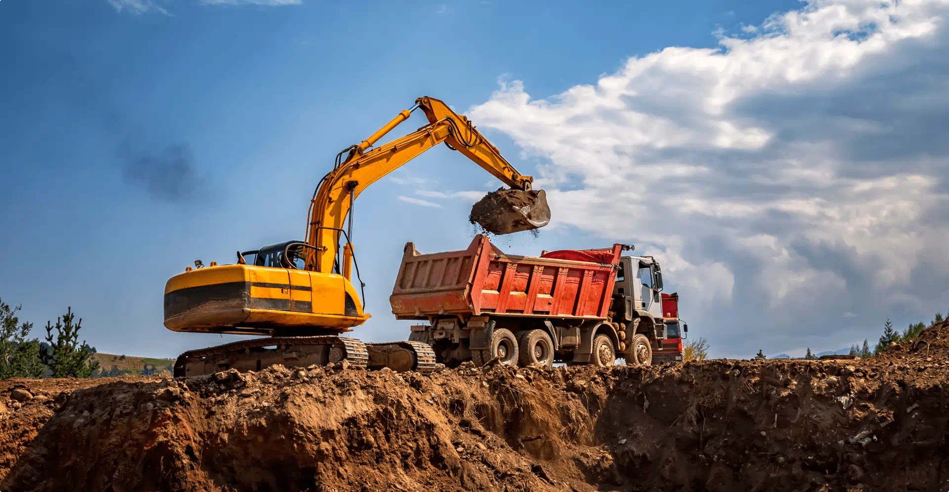 A yellow excavator lifts soil into a red dump truck at an open construction site, with a deep trench in the foreground and clouds overhead.