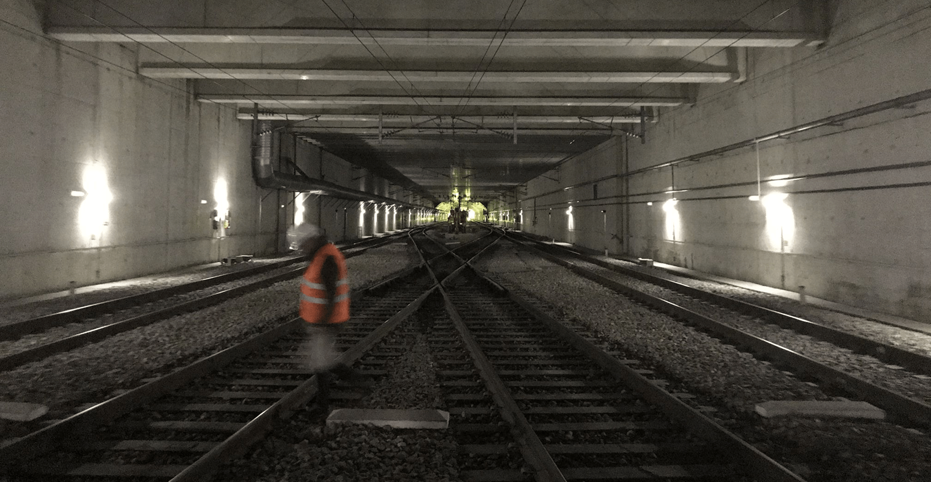 A man walks along train tracks inside a dimly lit tunnel, surrounded by concrete walls and shadows.