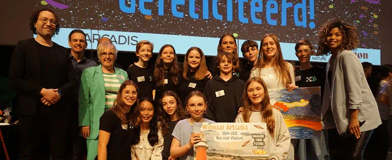 A group of people smiling and posing together for a photo on a stage, with a backdrop and audience visible behind them.