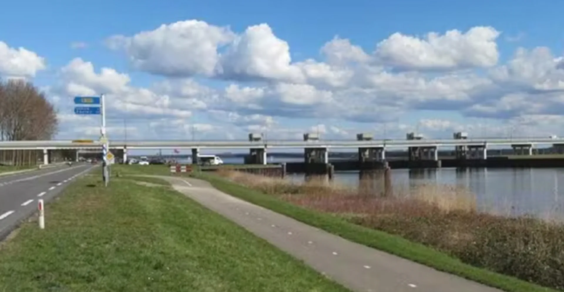 A bridge crosses a river, with a road visible on the far side, framed by trees and a bright sky.