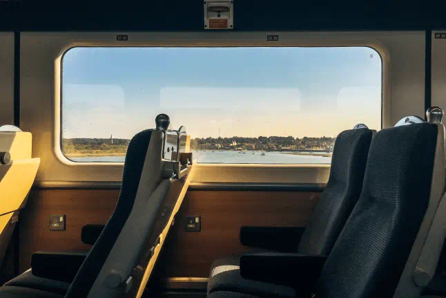Interior of a modern passenger train with comfortable blue seats and a large window overlooking a scenic coastal landscape, showcasing a relaxing rail travel experience during golden hour.