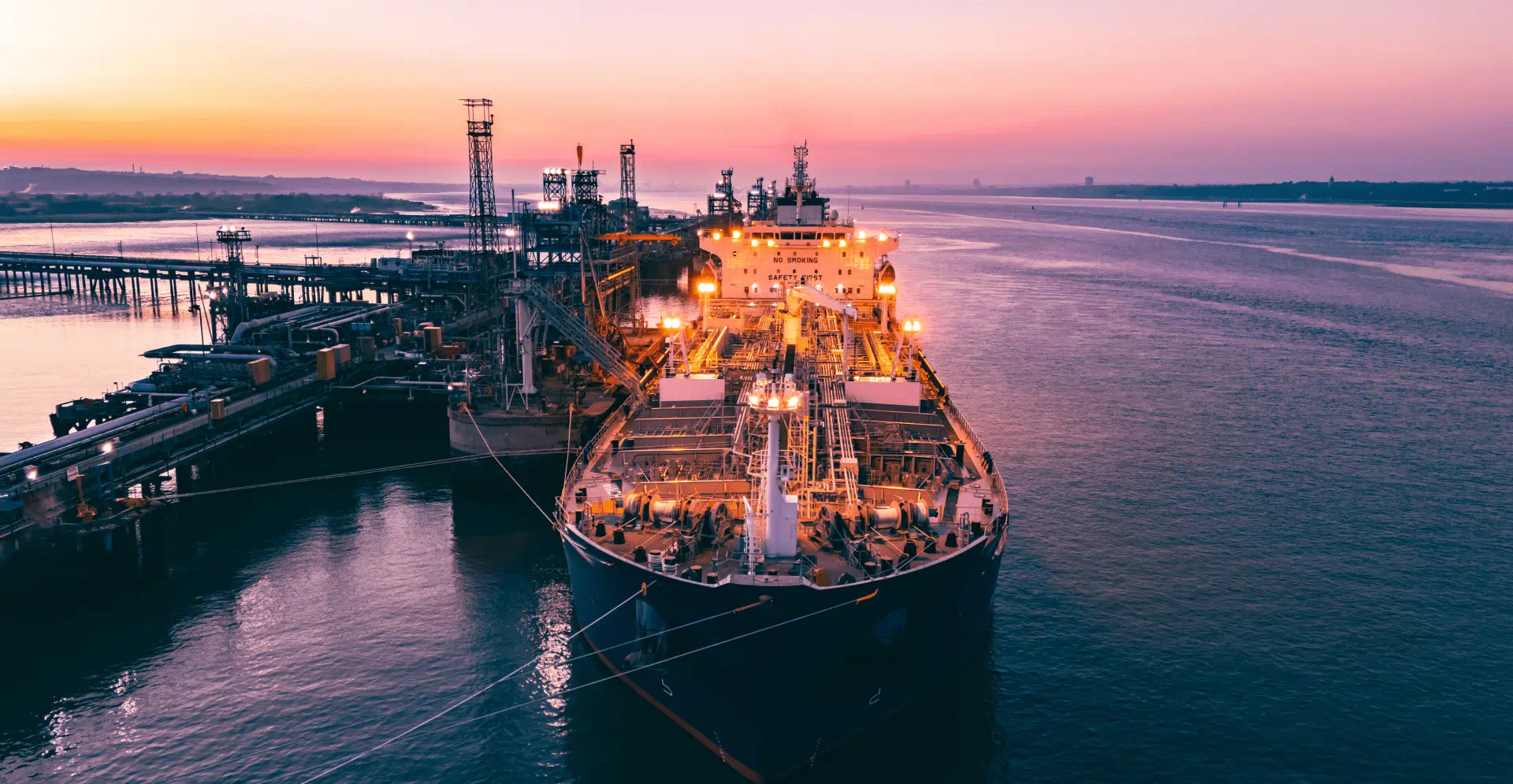 Aerial view of a large, illuminated cargo ship docked at an industrial port during sunset. The sky is a gradient of pink and purple, reflecting on the calm water.