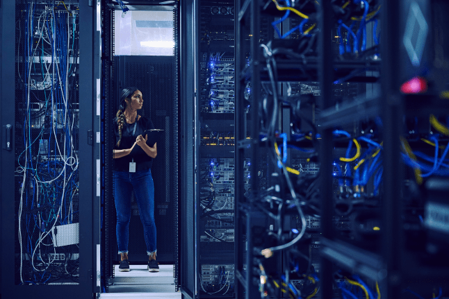 A woman in a server room inspects wires and servers, demonstrating her role in technology management.