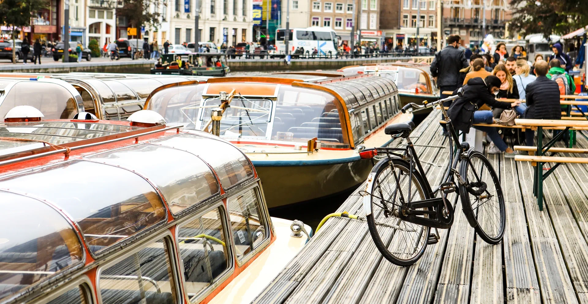 Canal landscape in Amsterdam with boats, a bicycle parked on a wooden on the dock with people on the background.