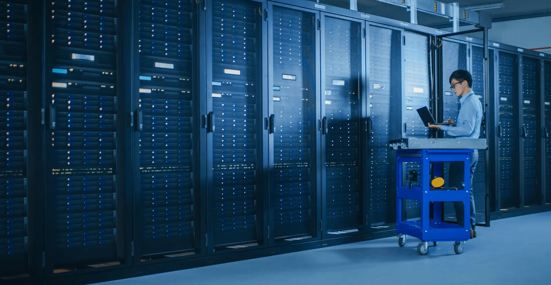  In Data Center: Male IT Technician Running Maintenance Programme on a Laptop, Controls Operational Server Rack Optimal Functioning. Modern High-Tech Telecommunications Operational Super Computer.