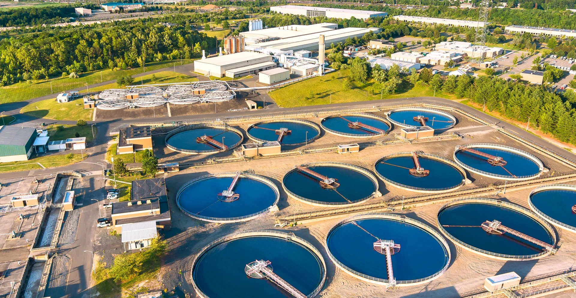 Aerial view of a wastewater treatment plant with multiple circular sedimentation tanks, surrounding buildings, and green landscape.