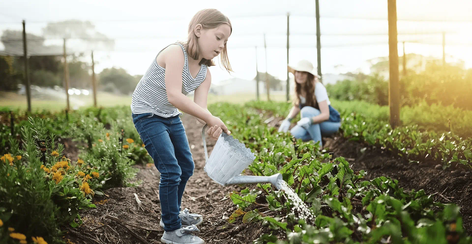 A young girl waters plants in an organic garden, surrounded by vibrant greenery and blooming flowers.