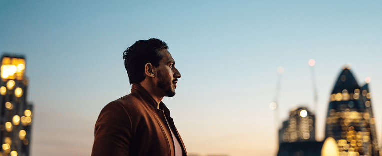 A man stands confidently in front of a vibrant city skyline, showcasing tall buildings against a clear blue sky.