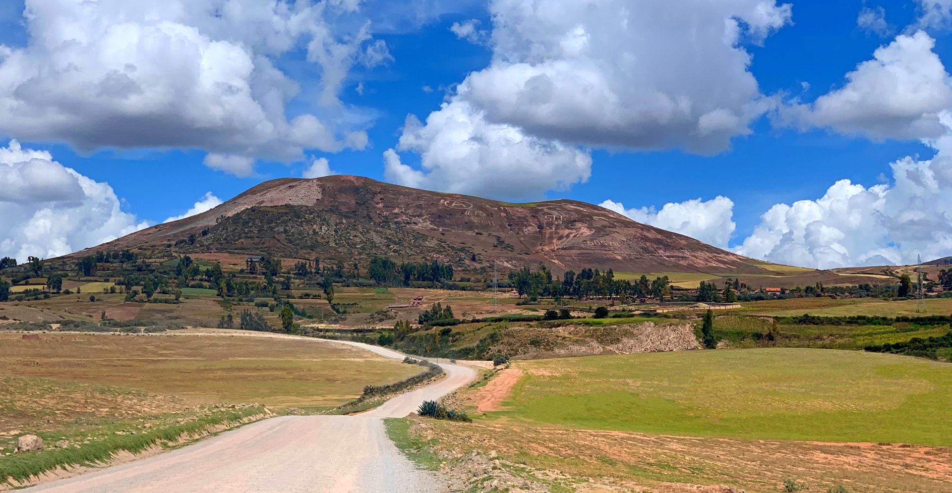 A rough road winds through a grassy field, leading to a mountain range in the background under a clear blue sky.