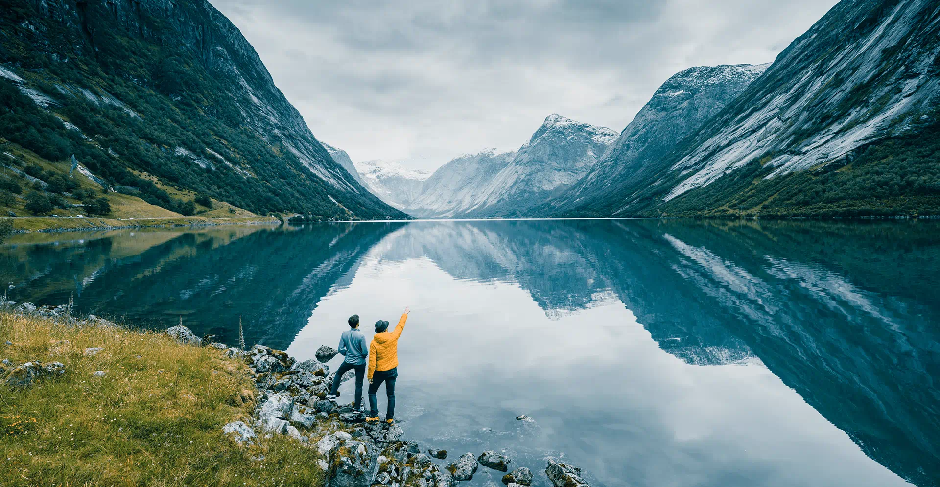 Two people stand on a lakeshore, surrounded by mountains, enjoying the serene landscape.