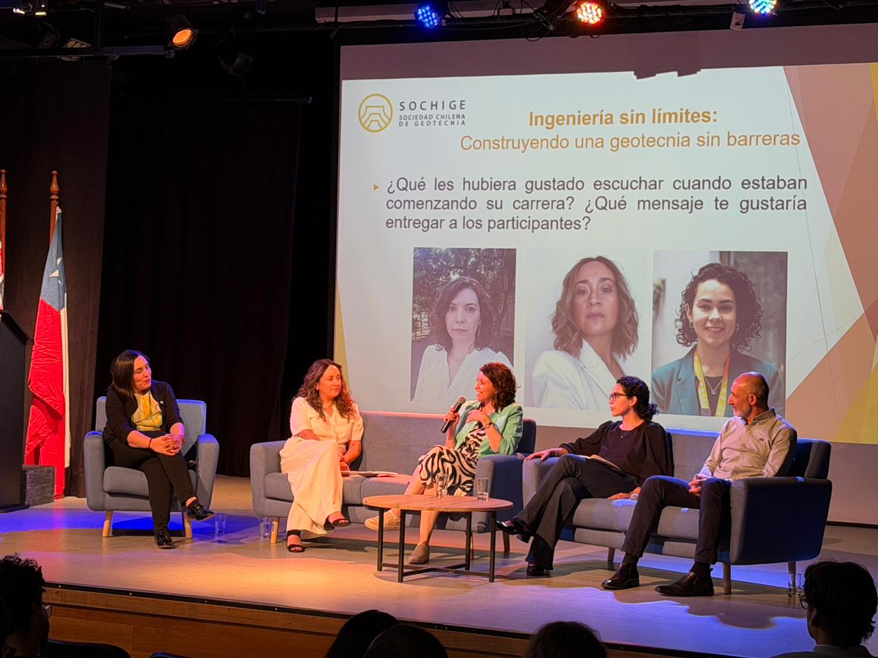 A group of people seated on a stage, engaged in a presentation, with a projector screen displaying visuals behind them.