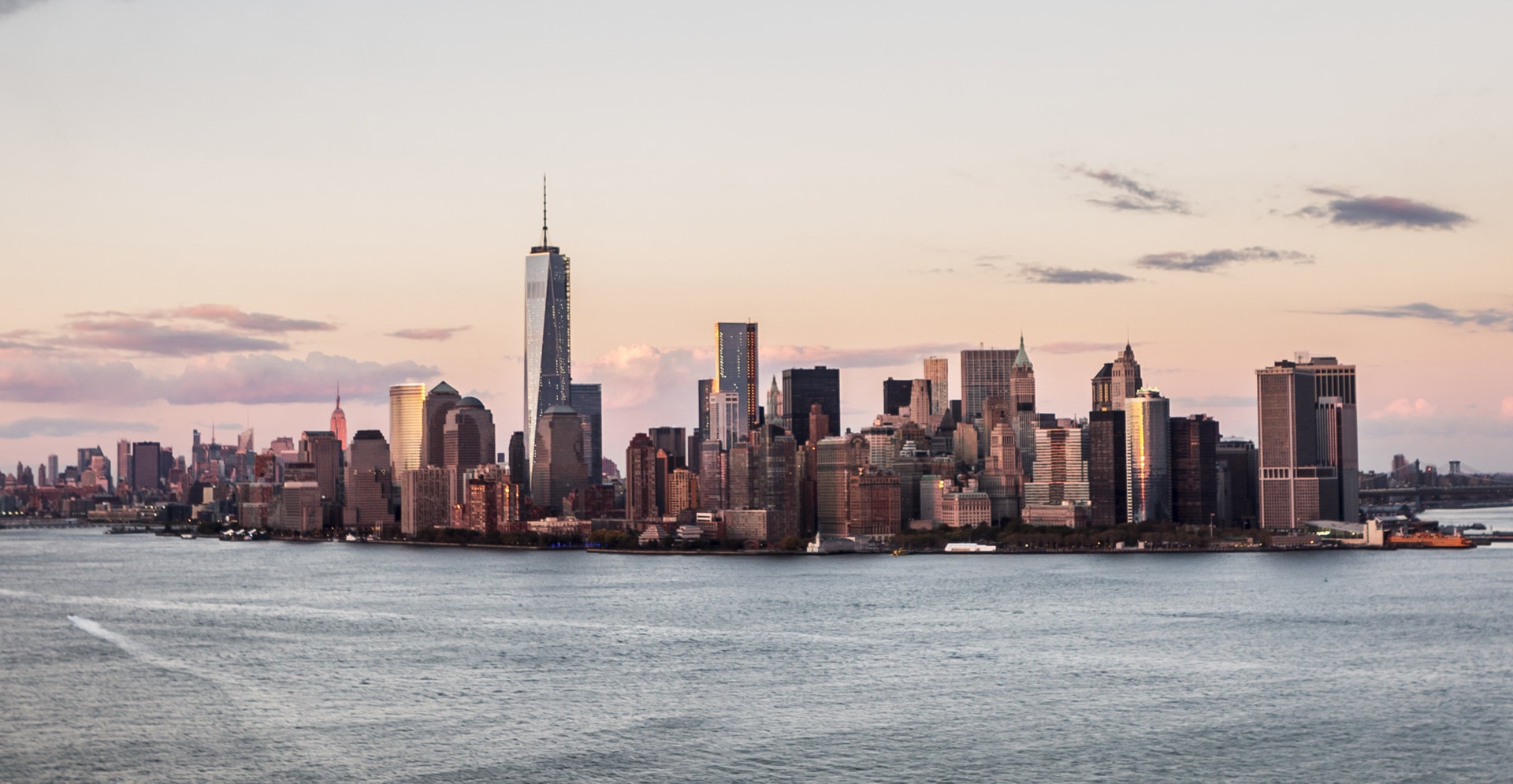 City skyline showcasing towering buildings and urban landscape over a calm lake with vibrant orange and pink colors reflecting on the water.