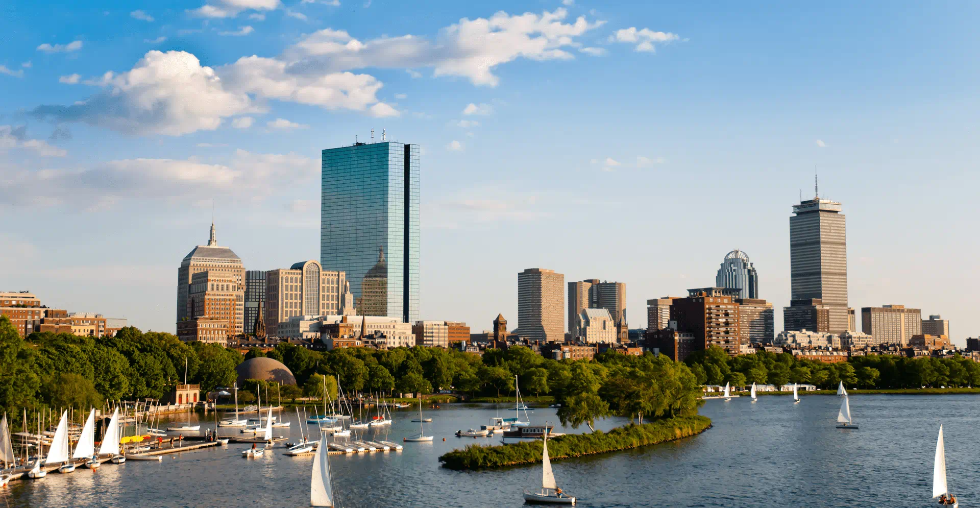 Boston skyline with sailboats on the Charles River under a clear blue sky.