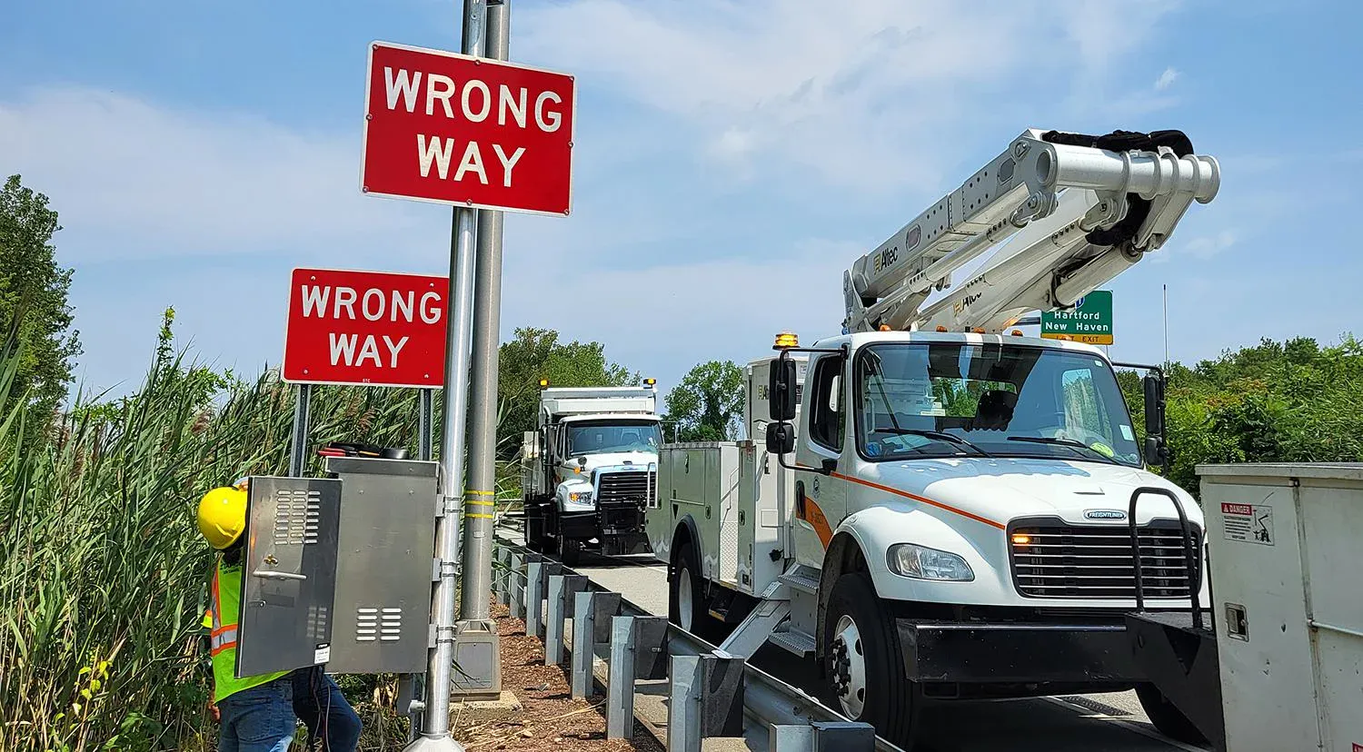 Utility trucks parked near "Wrong Way" signs on a road, with a worker in a hard hat inspecting equipment. The scene conveys caution and awareness.
