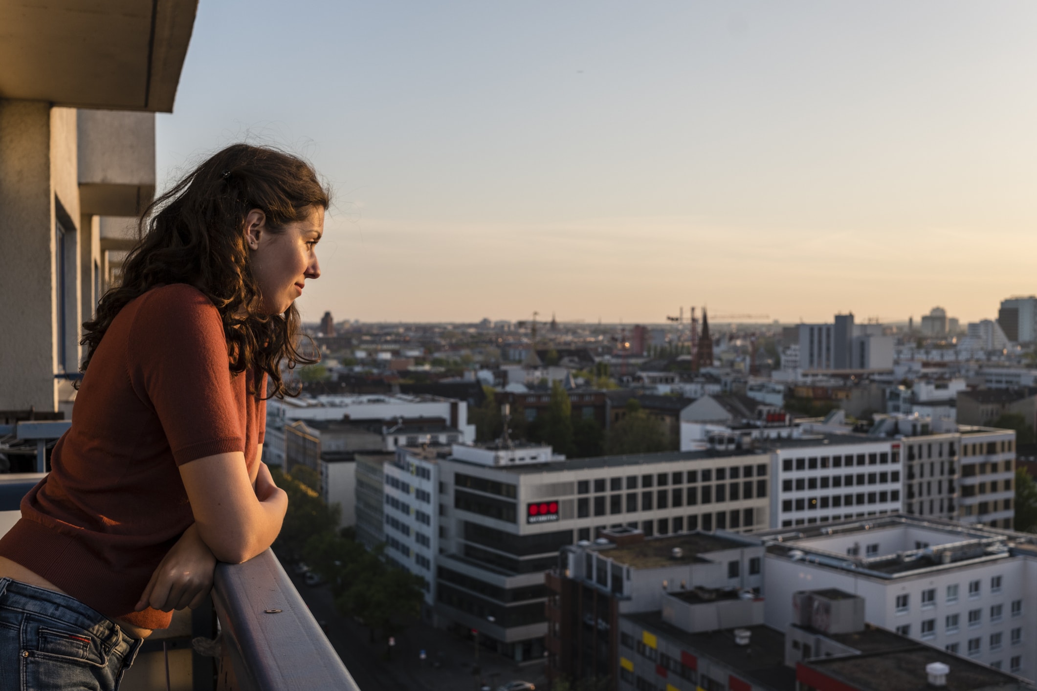 junge Frau lehnt sich auf Balkon und blickt bei Sonnenuntergang auf die Skyline von Berlin