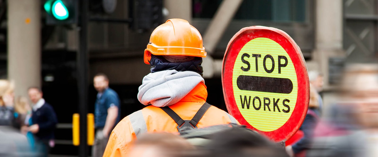 A man wearing an orange jacket and hard hat stands before a stop works sign, emphasizing safety in a construction area.