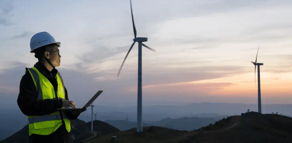 A man in a hard hat and vest standing in front of wind turbines at a renewable energy site.