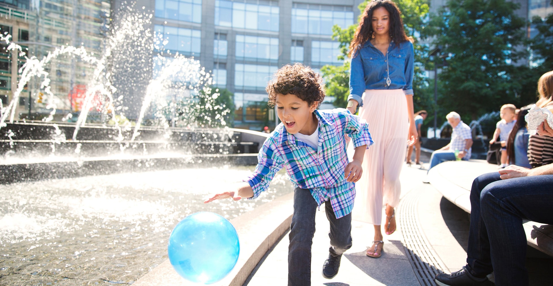 A child playing a ball, a woman walking and people gathered beside a fountain.