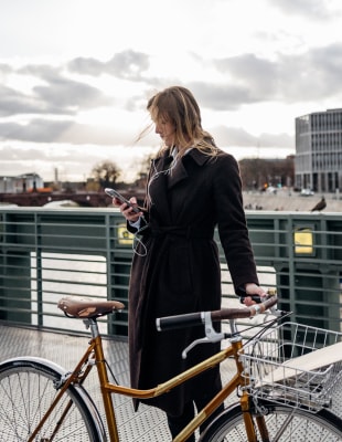 A woman in a coat stands beside a bicycle, showcasing a casual yet stylish outdoor look.